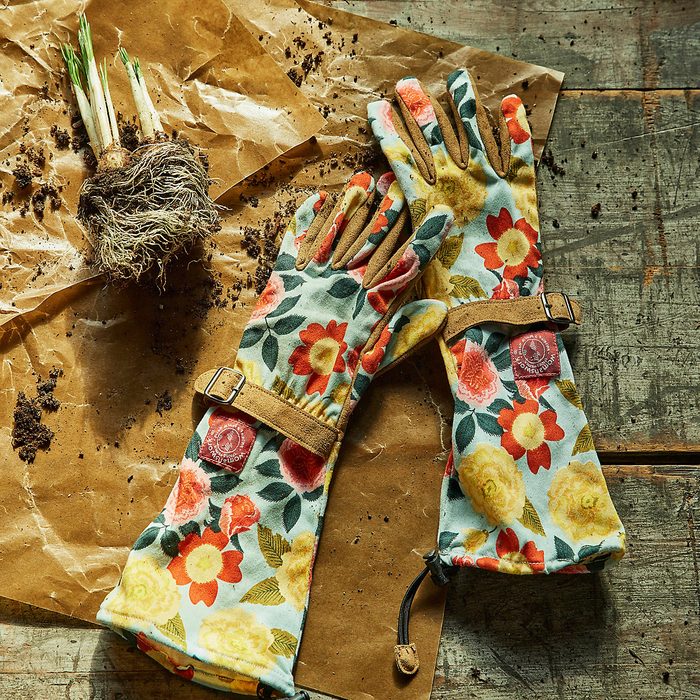 Floral gardening gloves rest beside a small bundle of roots and soil, arranged on crumpled brown paper atop a weathered wooden surface.
