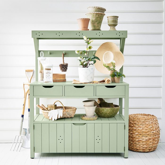 A green storage table holds gardening tools and pots, while a small plant and straw hat decorate its surface. The background features white vertical panels.
