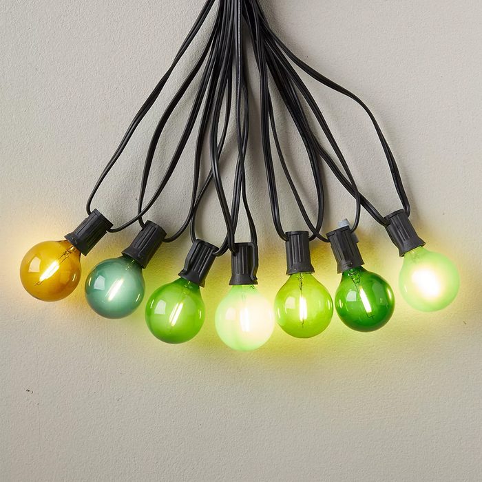 A string of eight light bulbs in various shades of green and yellow hangs against a plain wall, glowing softly along their length.