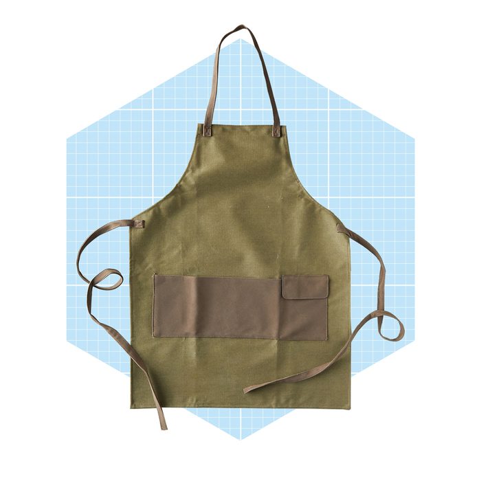 An olive green apron hangs against a light blue grid background, featuring straps and a pocket, suggesting readiness for cooking or crafting activities.