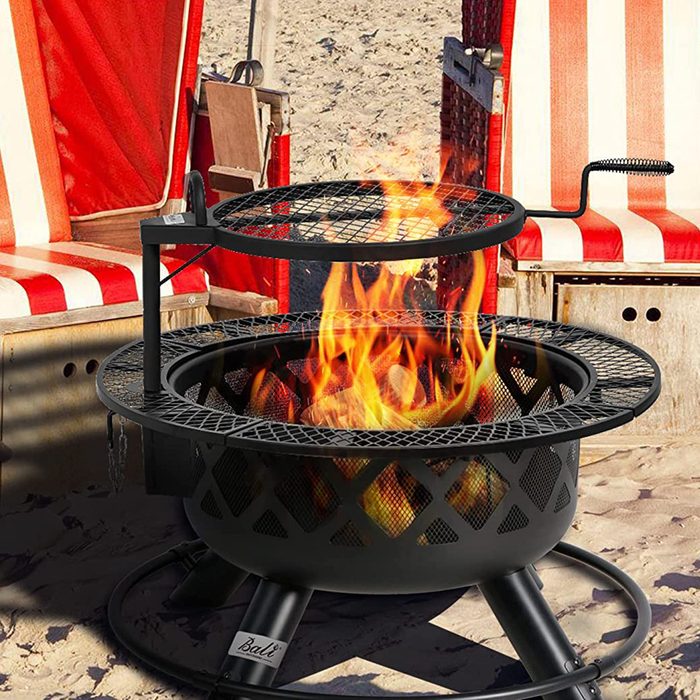 A black fire pit burns brightly, with flames visible through a mesh top, positioned on a sandy beach near red and white striped chairs.