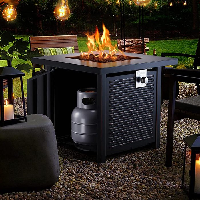 A square fire table has flames dancing above rocks, surrounded by outdoor seating and illuminated by hanging lights, creating a cozy atmosphere in a garden setting.