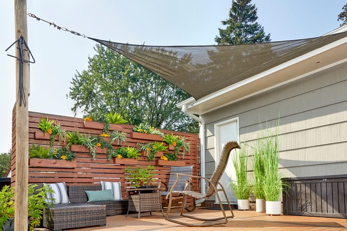 A shaded outdoor seating area features a rocking chair and wicker furniture, surrounded by a vertical garden and leafy plants against a house backdrop.
