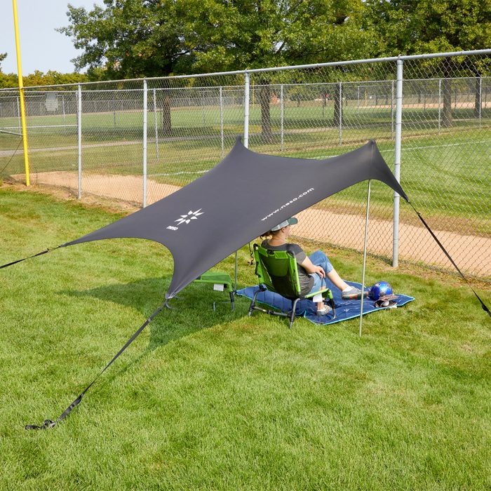 A person sits under a large black shade tent, relaxing in a green chair on grass near a diamond, surrounded by a chain-link fence and trees.