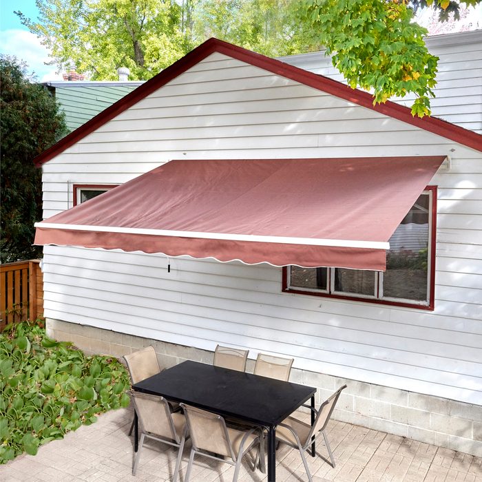 A brown awning extends over a black table surrounded by beige chairs, adjacent to a white house in a green, leafy outdoor setting.
