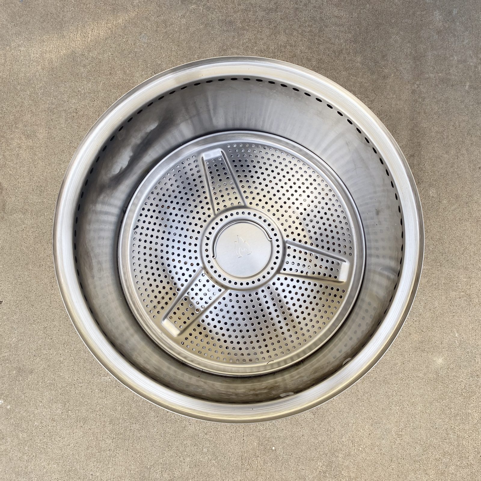A stainless steel pot with a perforated steamer insert sits on a flat, grey surface, ready for cooking or boiling food.