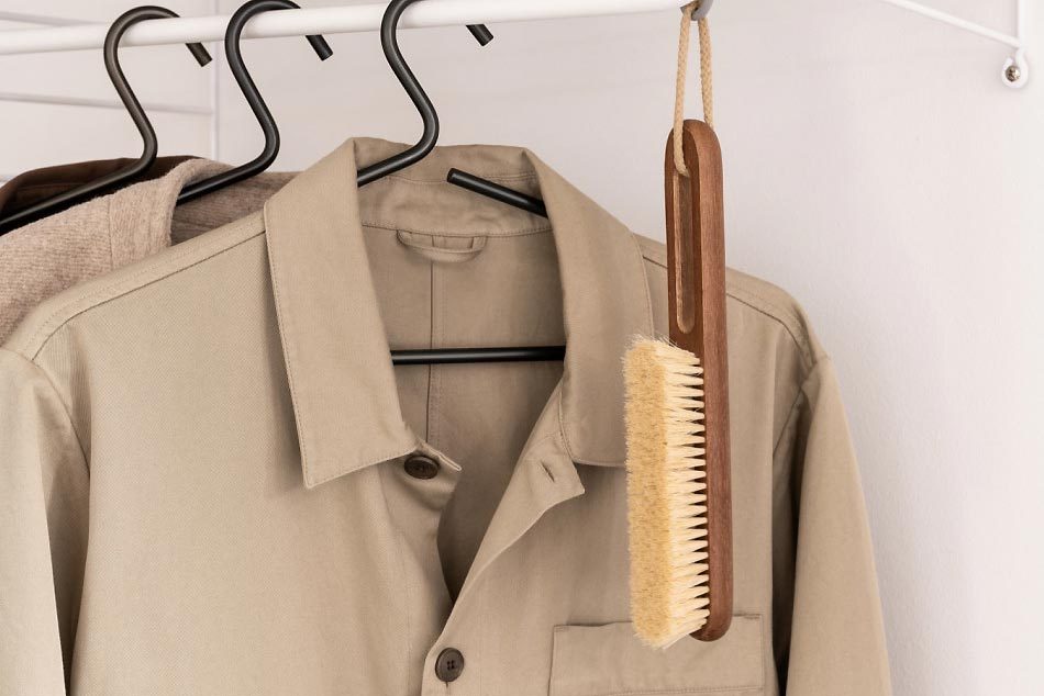 A beige shirt hangs on a white rack, with a wooden garment brush attached, resting against the fabric, in a minimalist interior setting.