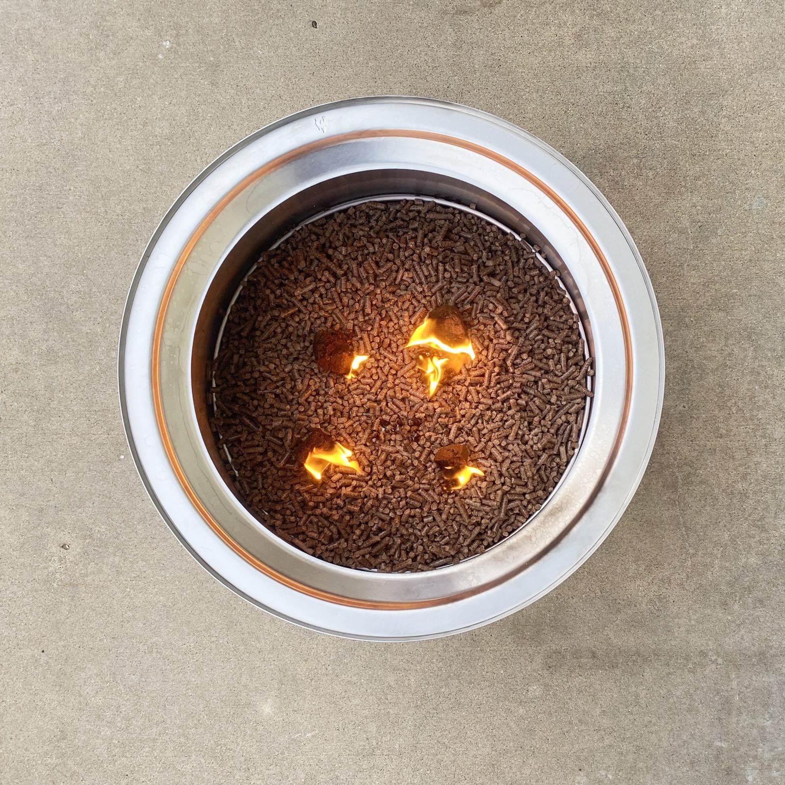 A metal container filled with small brown pellets has flames emerging from some, resting on a flat, light-colored surface.