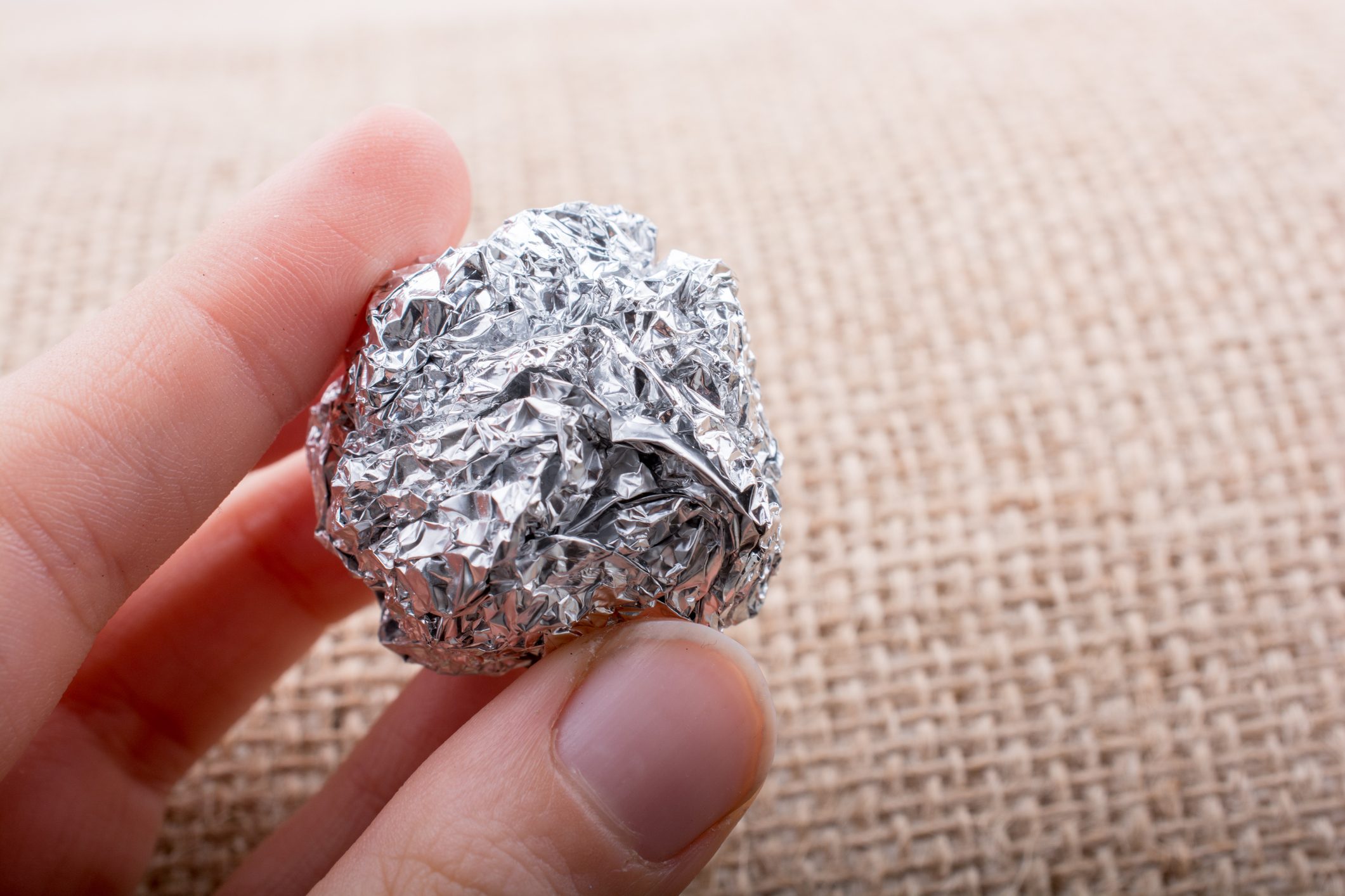 A hand holds a crumpled ball of aluminum foil above a textured, natural fabric surface.