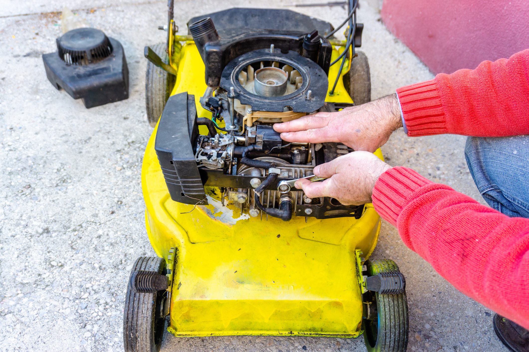 A person repairs the engine of a yellow lawn mower on concrete. Tools are placed nearby, and the scene is outdoors in a casual environment.