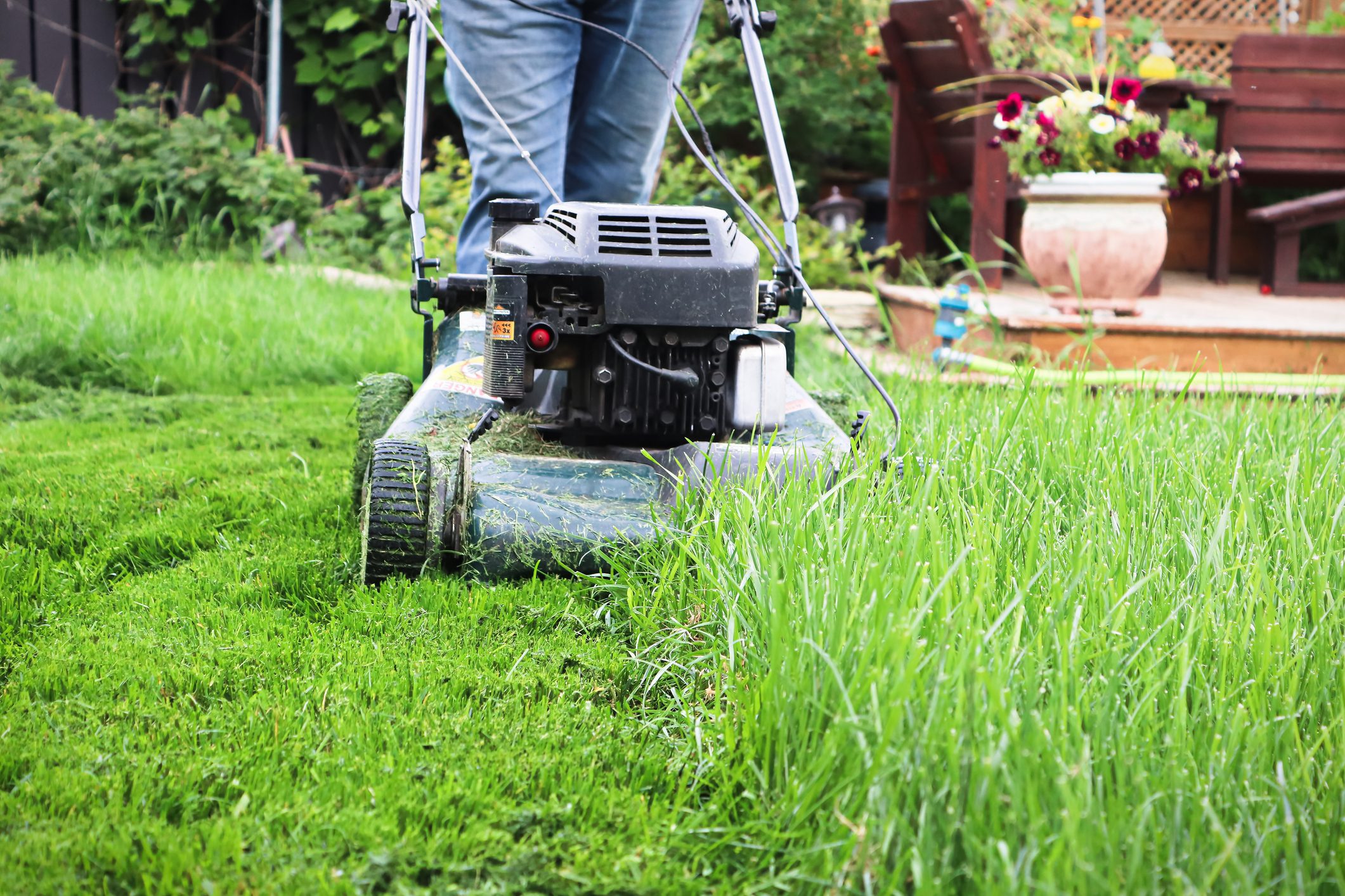 A lawnmower operates on overgrown grass, trimming it evenly, while a garden space with planters and seating is visible in the background.