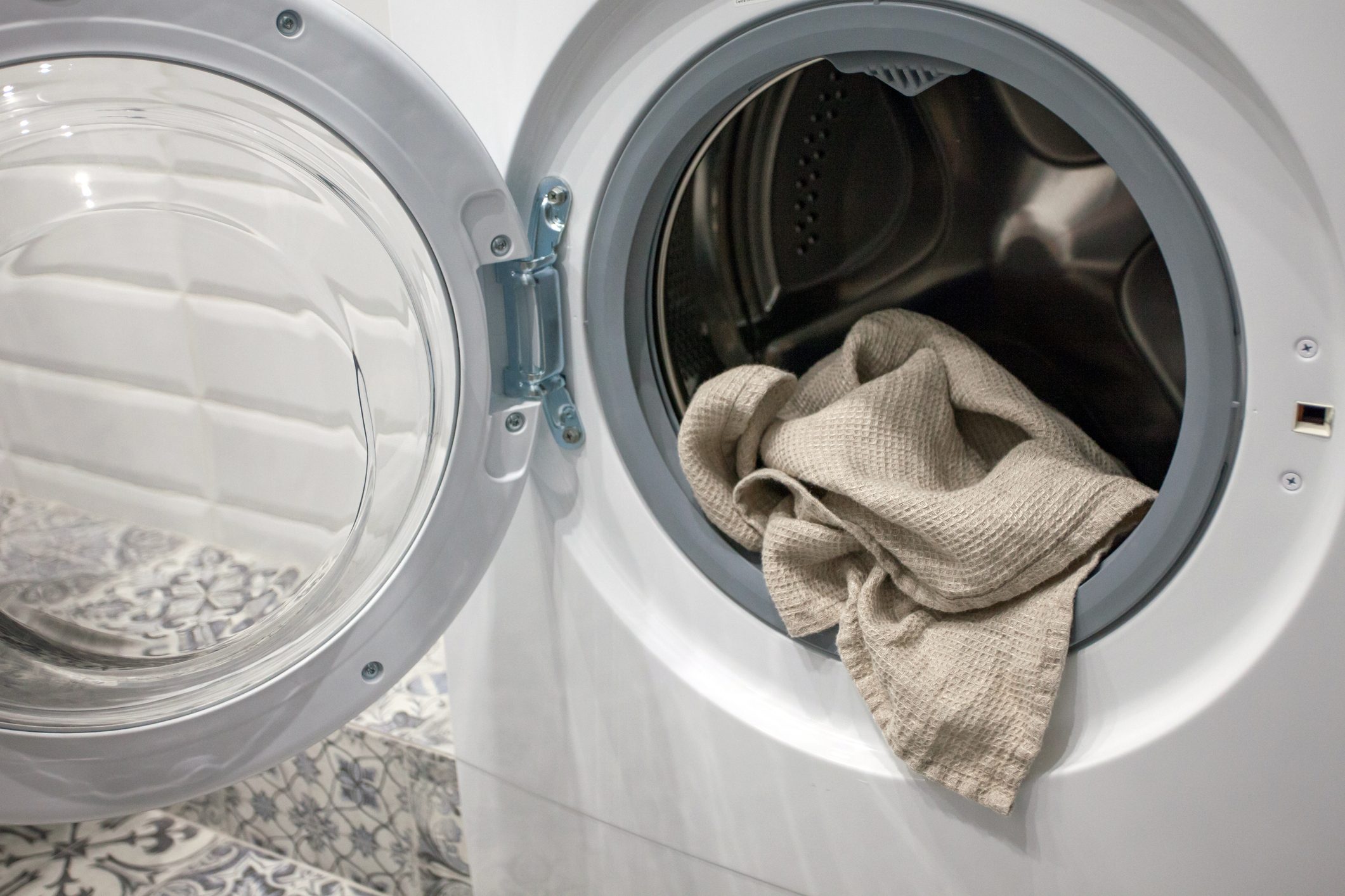 A partially open washing machine door reveals a crumpled beige towel inside, set against a patterned tiled floor in a modern laundry space.