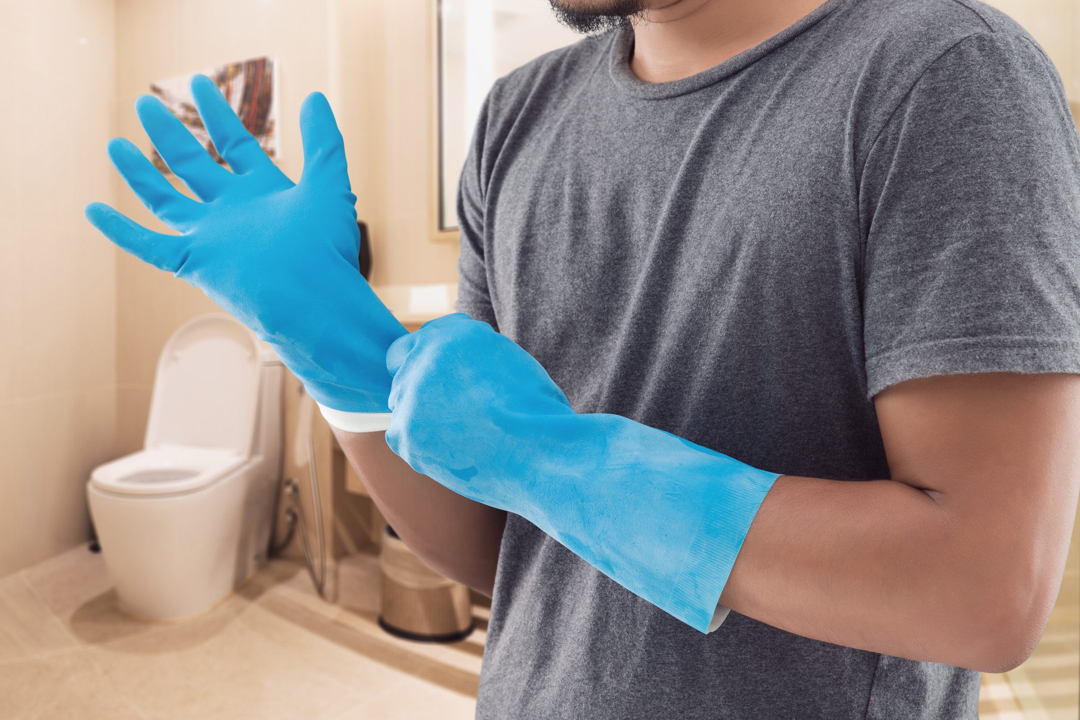 A person puts on blue rubber gloves while standing in a bathroom, with a toilet visible in the background.