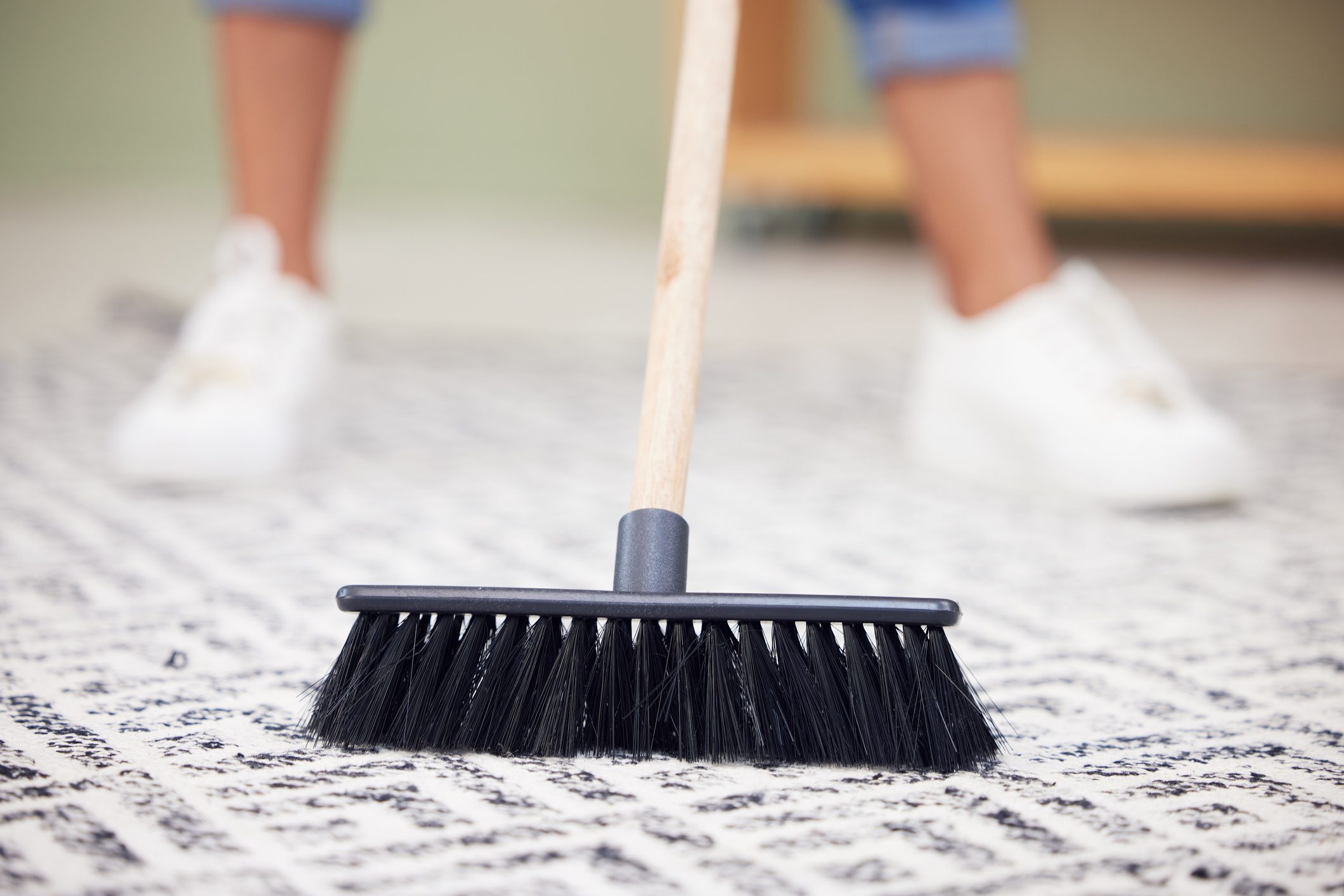 A broom sweeps across a patterned rug as a person in casual clothing stands nearby, contributing to cleaning in a light, indoor space.