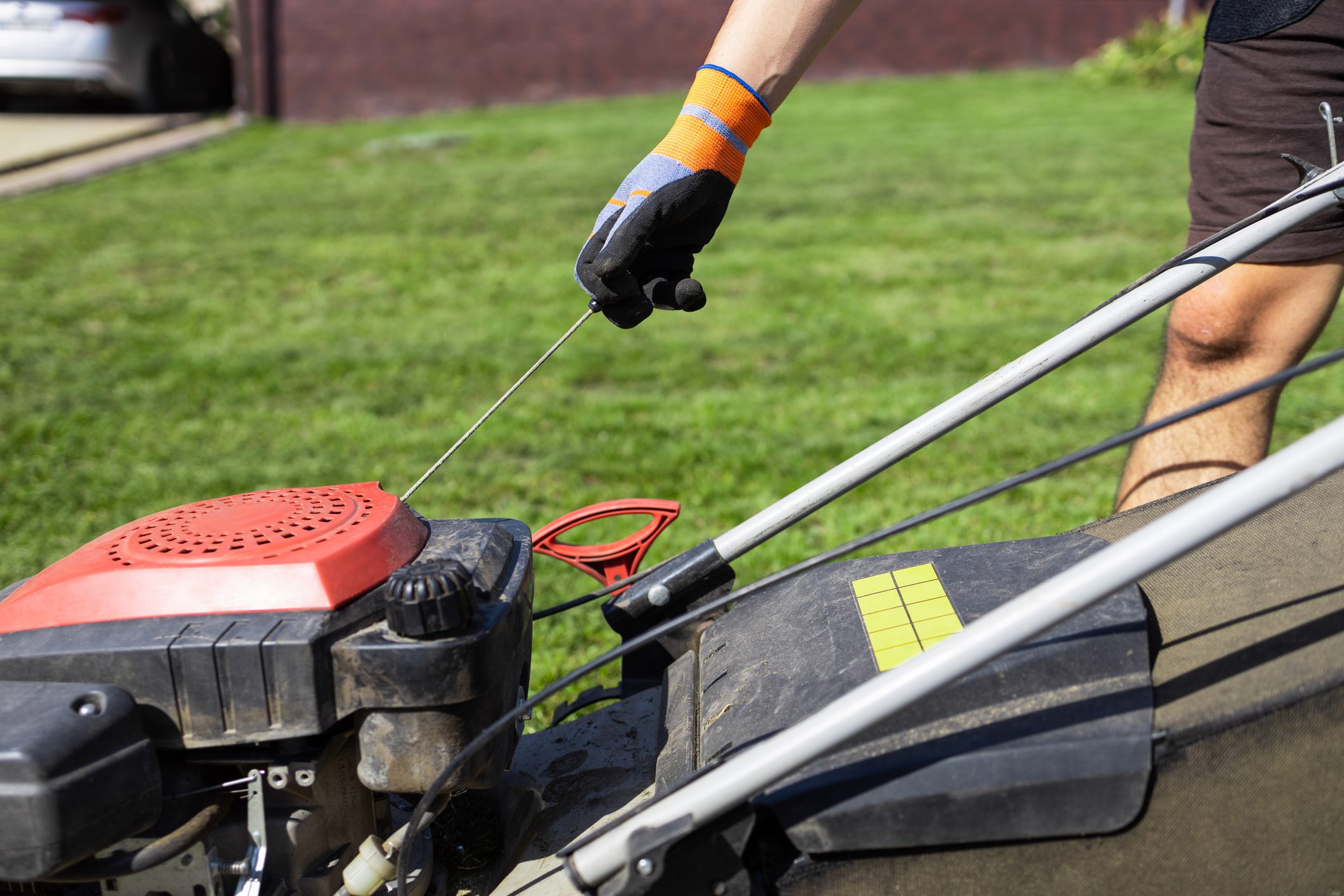 A gloved hand pulls the starter cord of a lawnmower situated on a manicured lawn, with a vehicle visible in the background.