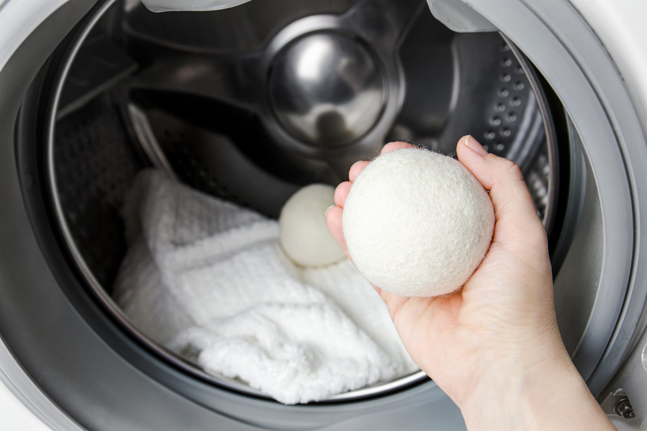 A hand holds a white felted laundry ball, positioned above a washing machine drum containing a folded white towel and another ball.