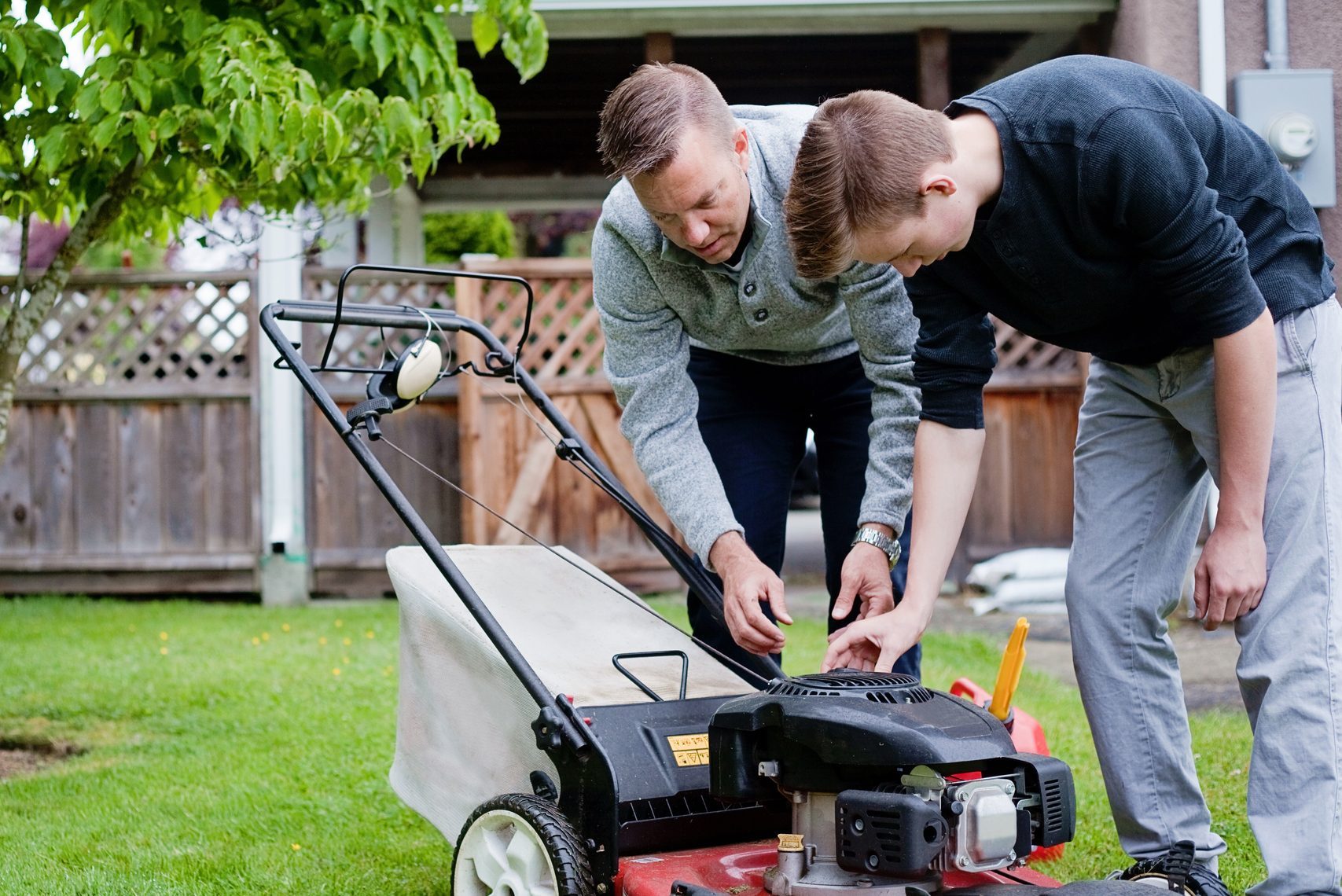 Two individuals examine the engine of a lawn mower on a grassy yard, surrounded by trees and a wooden fence.