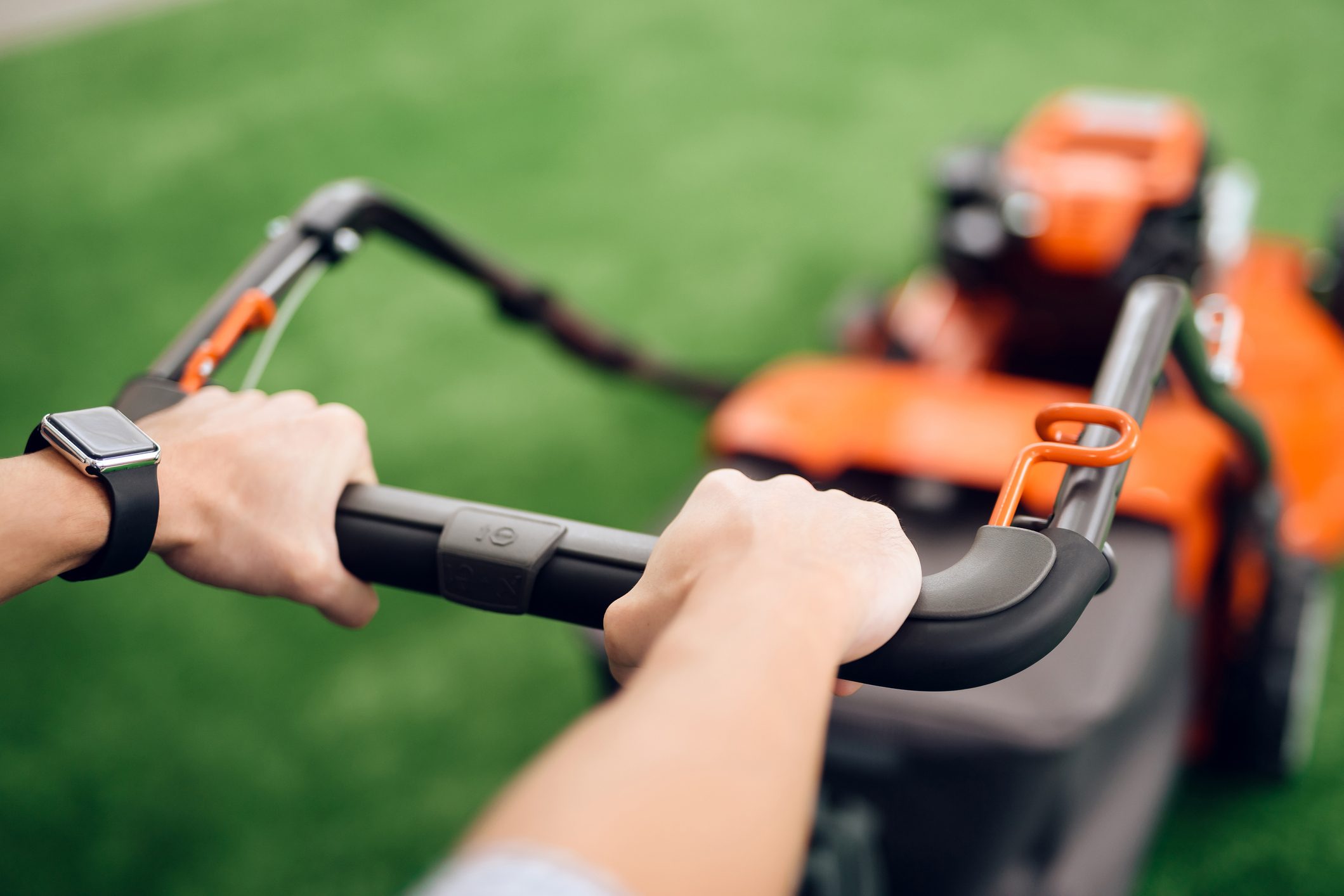 A hand grips a lawnmower's handle, navigating across a manicured green lawn, ready to cut the grass. A smartwatch is visible on the wrist.