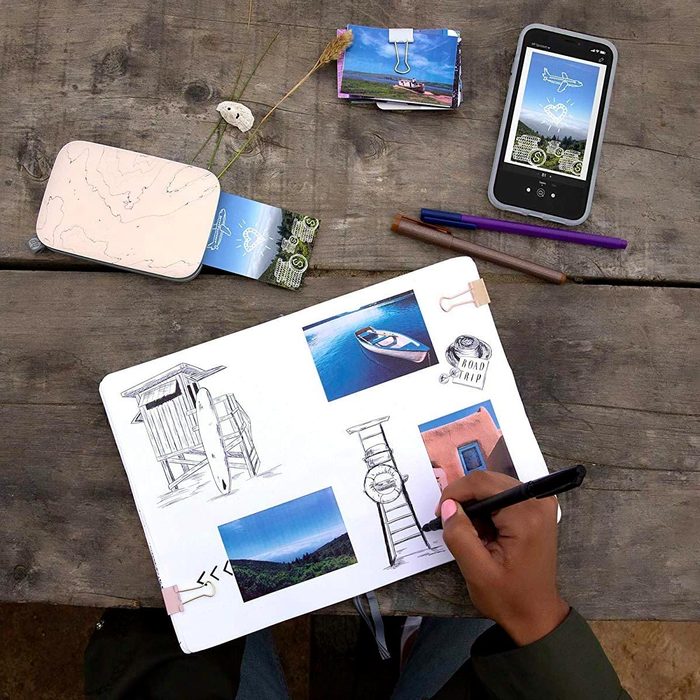 A hand holds a pen, drawing on a sketchbook adorned with photos, while a smartphone and scattered images lie on a wooden table.