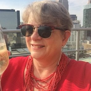 A woman in sunglasses smiles while holding a glass of champagne. She wears a red shirt and necklace, surrounded by a city skyline on a sunny day.