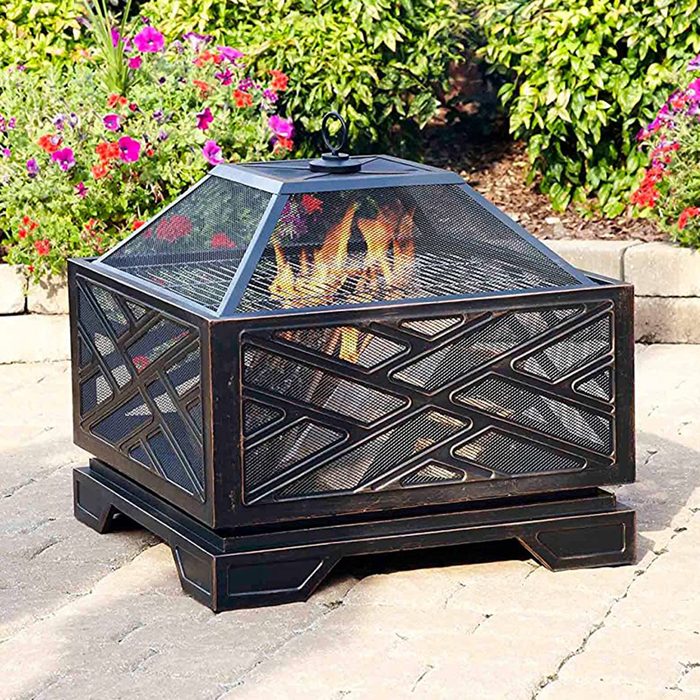 A square fire pit with a geometric metal design has flames burning inside, placed on a stone patio surrounded by flowering plants and greenery.