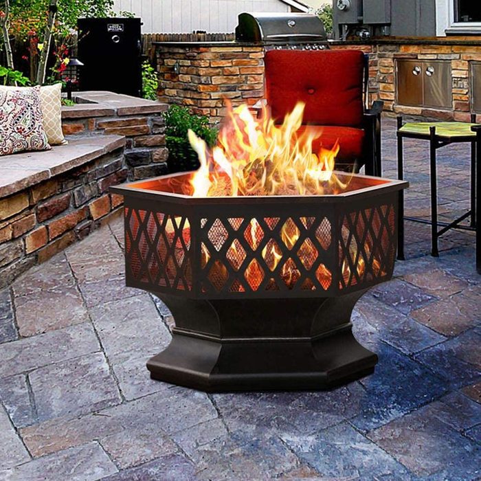 A metal fire pit emits flames, providing warmth in a stone-paved outdoor setting with seating and a grill nearby.
