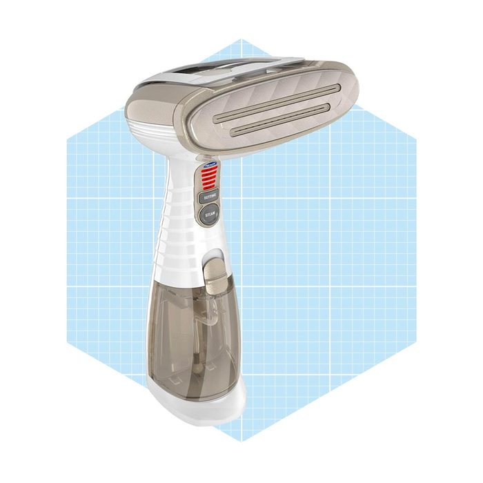 A handheld garment steamer is positioned upright, ready to emit steam, against a light blue grid background, indicating its cleaning purpose in fabric care.