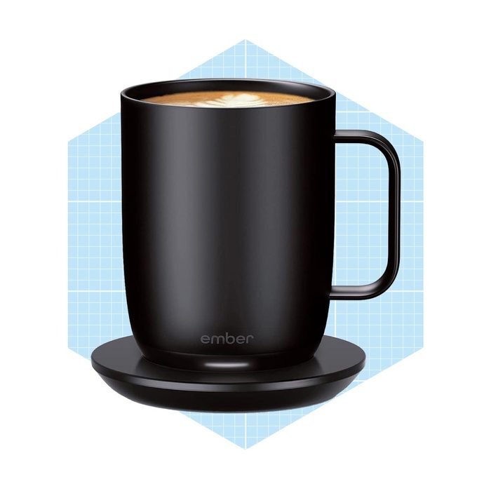 A black mug with a handle sits on a round heating coaster, containing a frothy beverage, against a light blue grid background.