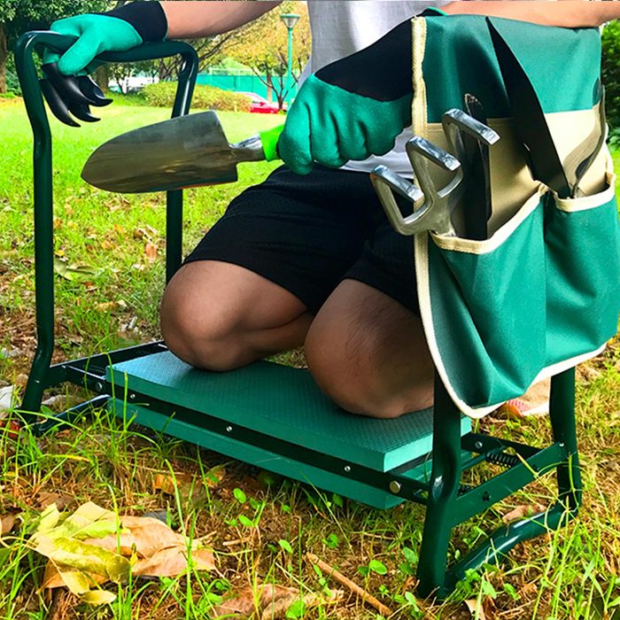 A person kneels on a green garden kneeler, holding a trowel and surrounded by gardening tools in a bag on the kneeler, amidst grass and leaves.