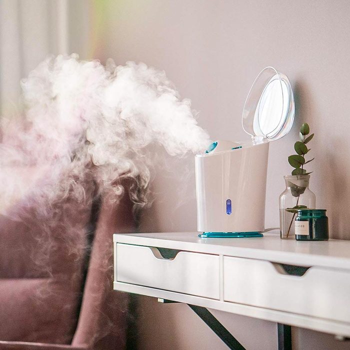 A humidifier releases mist into the air, set on a table near a plant and a candle, creating a calming atmosphere in a cozy room.