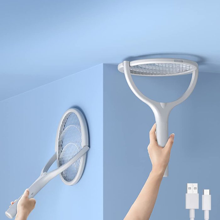 A hand holds a tennis racket-like device against a blue wall, while another similar device is mounted on the wall, suggesting usage for insect removal.