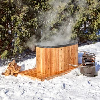 A wooden hot tub steams in a snowy outdoor setting, accompanied by a stacked firewood pile and a metal heating apparatus beside it.