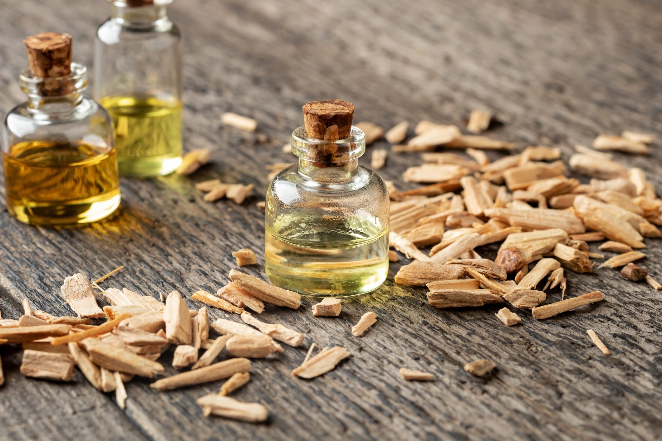 Glass bottles of oil are placed beside scattered wooden shavings on a textured wooden surface, creating a natural and rustic arrangement.