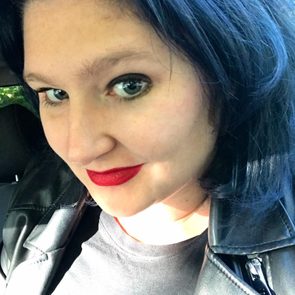 A woman with black hair and a red lip smile sits in a vehicle, illuminated by soft sunlight, wearing a black top and leather jacket.