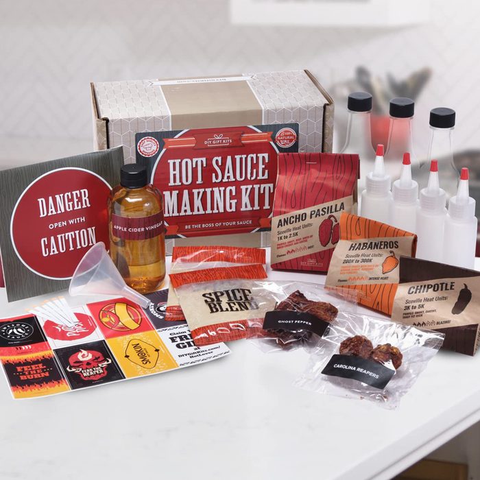 A hot sauce making kit includes various ingredients and tools, such as vinegars, peppers, spice blends, and bottles, arranged on a light surface.