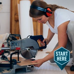 A woman uses a power saw to cut wood on a work surface, focused on her task in a partially furnished, well-lit room. Text: