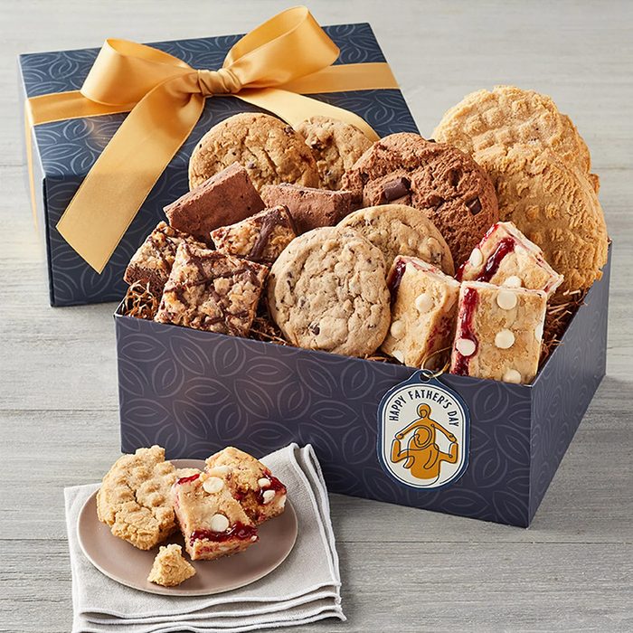 A gift box filled with assorted cookies and bars sits on a table, with a plate displaying two cookies and a slice beside it.