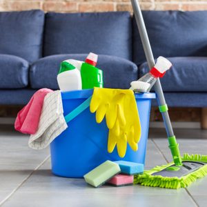 A blue cleaning bucket filled with supplies, including sponges, gloves, and bottles, stands on tiled flooring in a room with a dark couch nearby.
