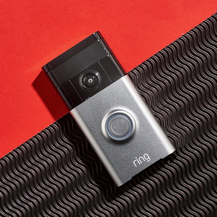 A video doorbell rests on a textured surface, capturing its sleek design. The background features a vibrant red color, contrasting with the doorbell