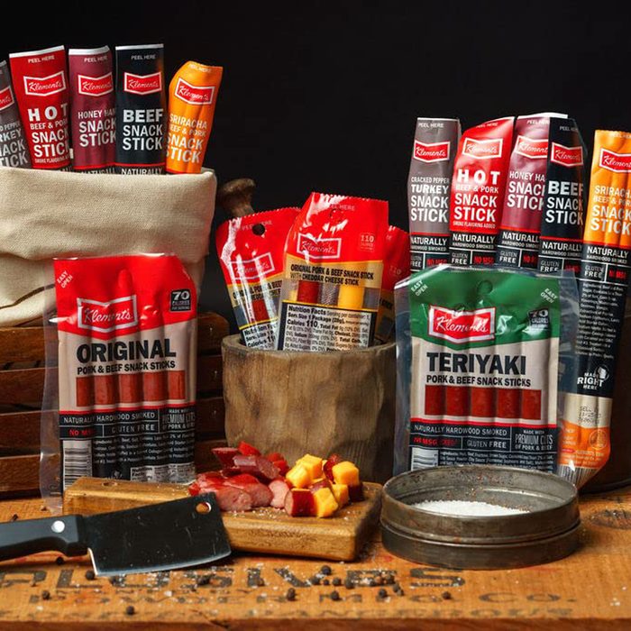 A variety of snack sticks, including beef and pork flavors, are displayed with a cutting board, knife, and cheese chunks on a rustic wooden surface.
