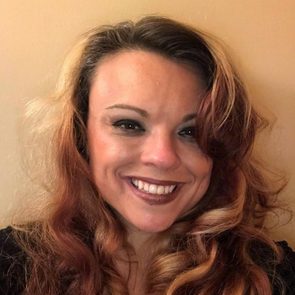 A woman with wavy, reddish-brown hair smiles broadly, showcasing her teeth. The background is a plain, light-colored wall, creating a warm, inviting atmosphere.