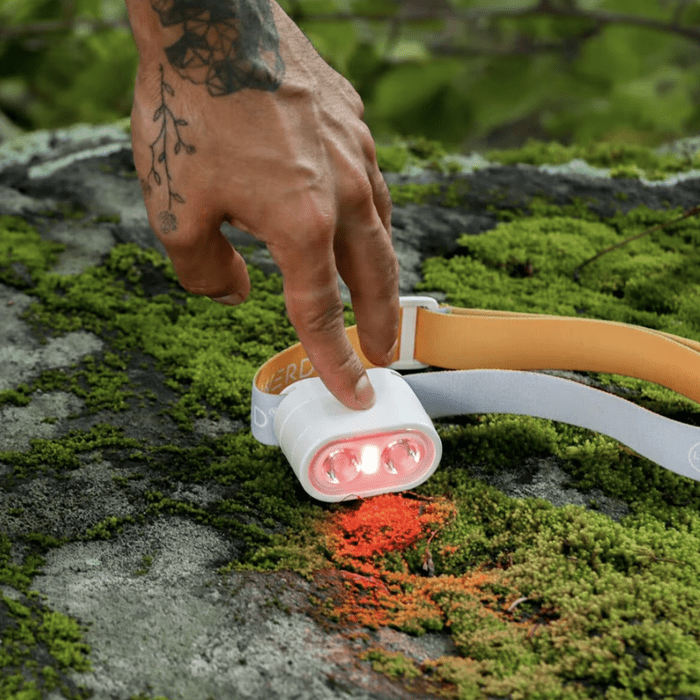 A hand touches a headlamp on a mossy rock, activating its lights in a natural outdoor setting. Bright colors contrast with the greenery.