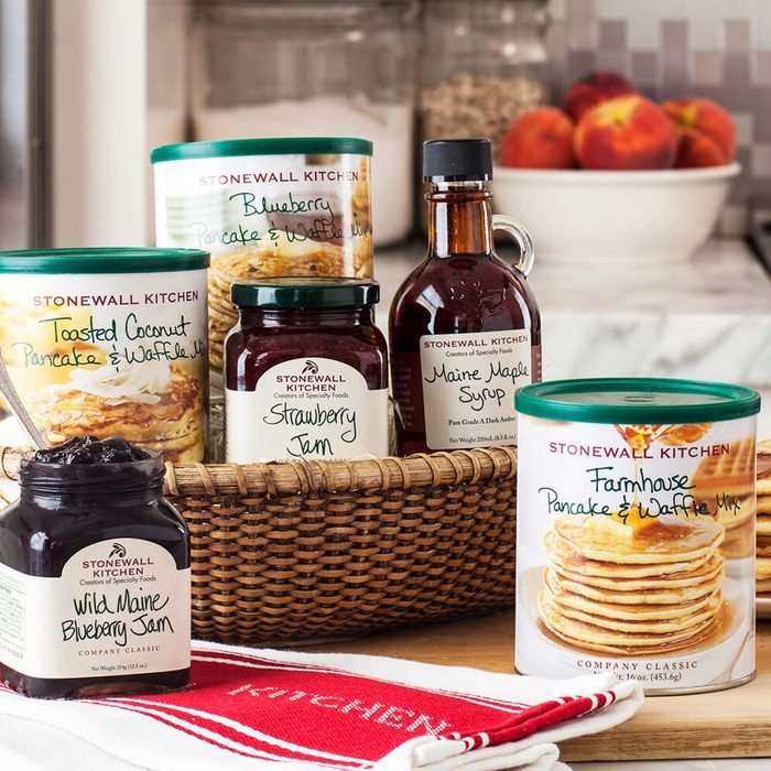 A basket holds various pancake mixes and jams, with a bottle of maple syrup beside it, set against a kitchen backdrop with fruit.
