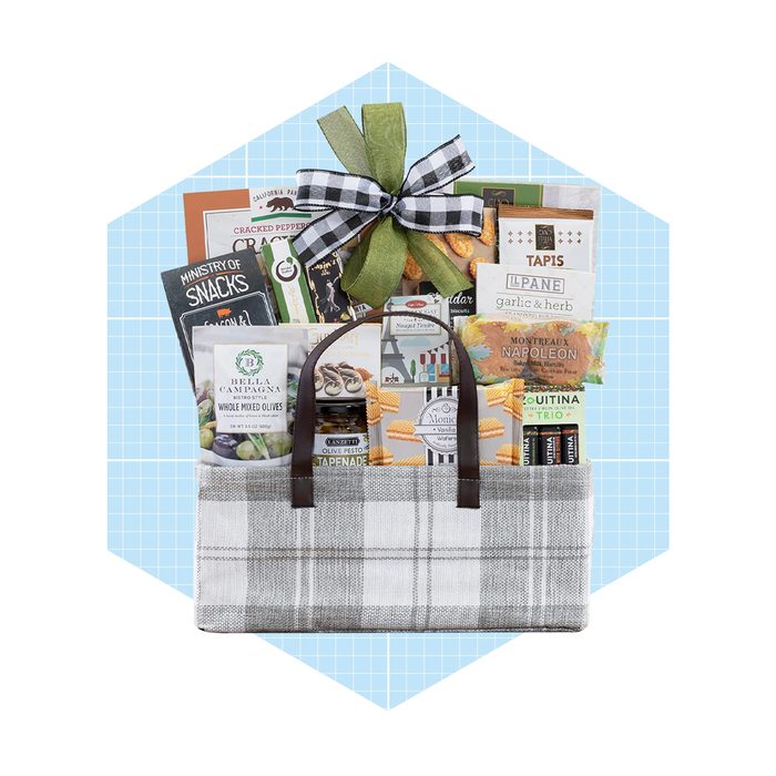 A checked fabric basket holds gourmet snacks, decorated with a green and black bow, set against a light blue geometric background.