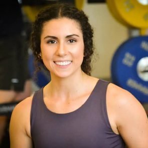 A smiling woman with curly hair poses in a gym setting, surrounded by weightlifting equipment, conveying a sense of confidence and fitness enthusiasm.