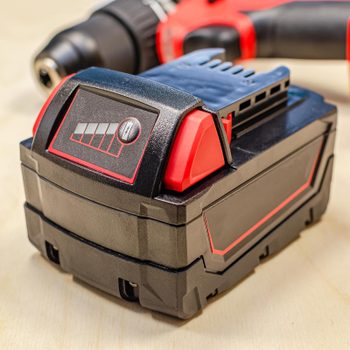 A battery sits on a wooden surface, displaying charge indicators, while an electric drill with a red and black design is partially visible nearby.
