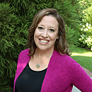 A woman with shoulder-length hair wears a black shirt and a purple cardigan, smiling while standing amidst green foliage in a well-lit outdoor setting.