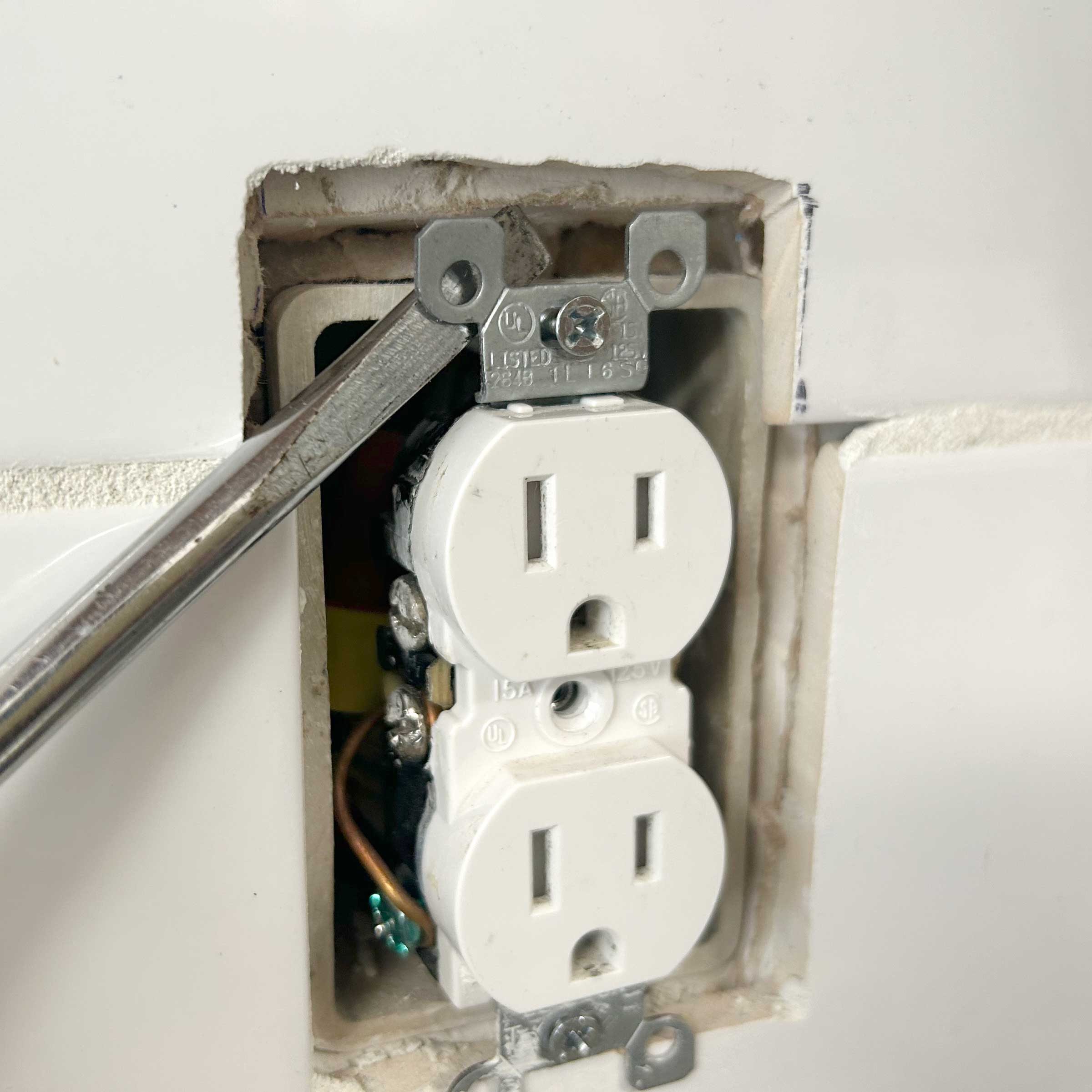 A white electrical outlet is being tightened with a wrench, situated in a partially exposed wall cavity. The background features drywall and unfinished edges.