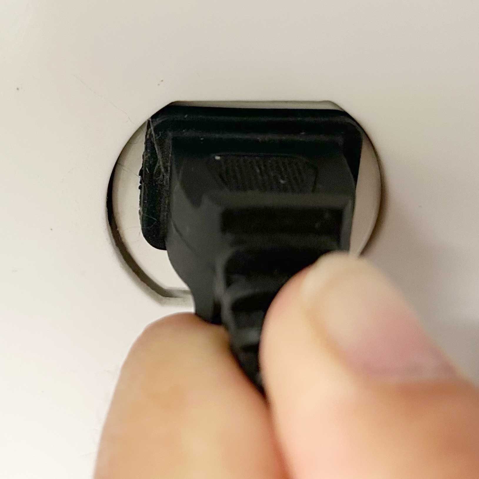 A hand grasps a black plug, inserting it into a circular electrical outlet on a white surface, indicating an action of connecting a device.