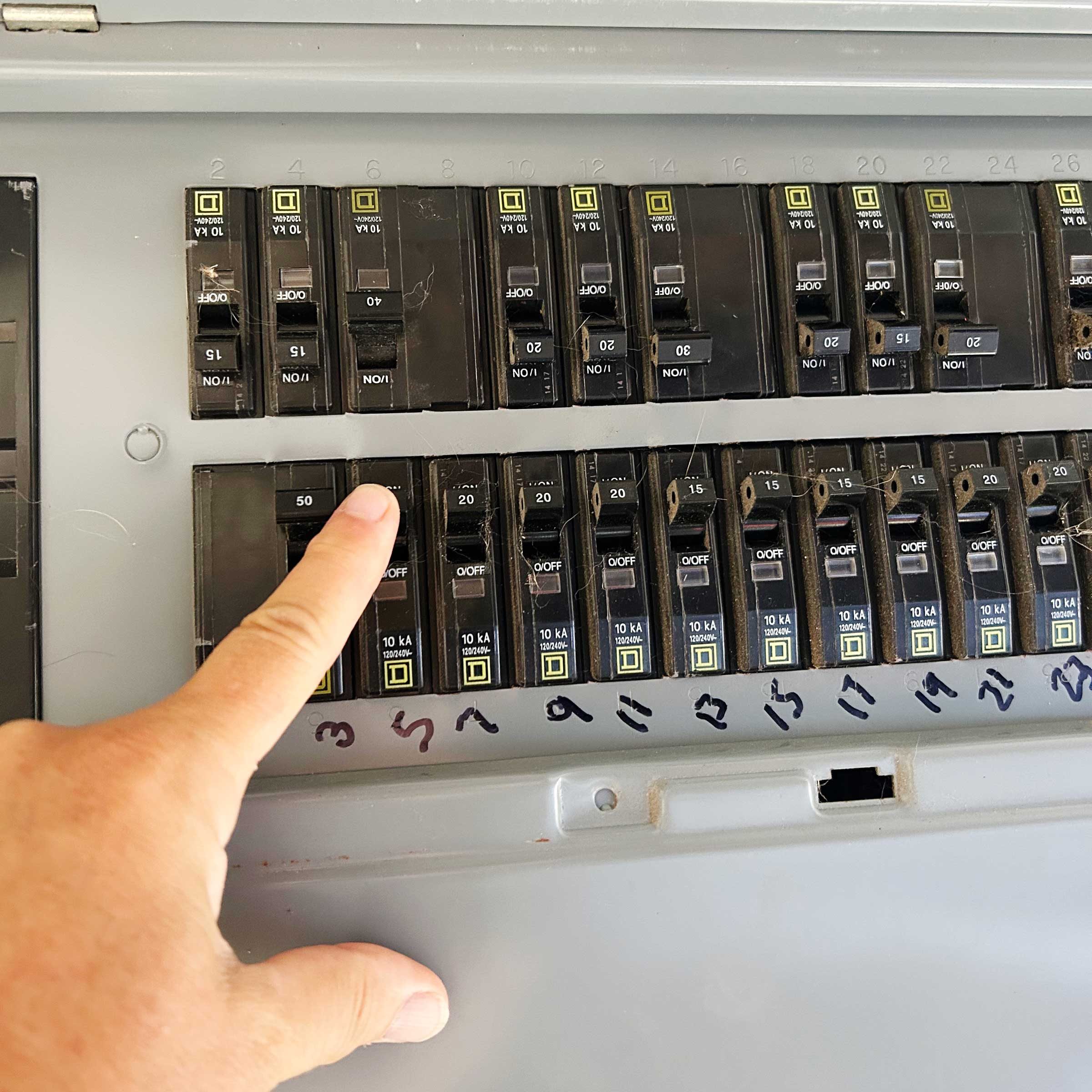 A hand presses a circuit breaker labeled "50" in a circuit panel filled with various switches, indicating maintenance or troubleshooting is taking place.